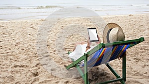 A woman holding a black tablet pc with blank desktop screen while lying down on a beach chair