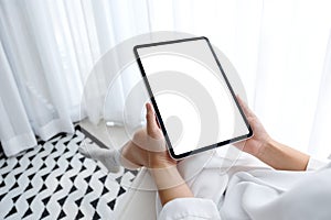 A woman holding black tablet pc with blank white desktop screen while sitting in bedroom with feeling relaxed in