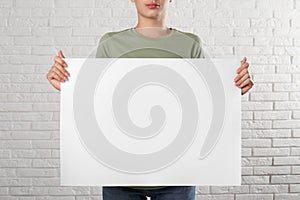 Woman holding blank poster near white brick wall, closeup. Mockup for design