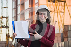 Woman holding blueprints, clipboard. Smiling architect in helmet at building