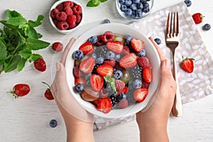 Woman holding bowl with fresh  fruit salad over white wooden table, top view