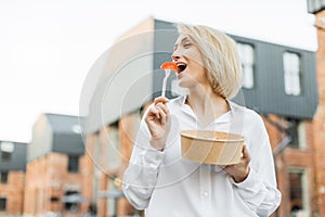 Woman holding a bowl of fresh salad in one hand and biting a slice of tomato on a fork in the other.