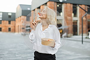 Woman holding a bowl of fresh salad in one hand and biting a slice of tomato on a fork in the other.