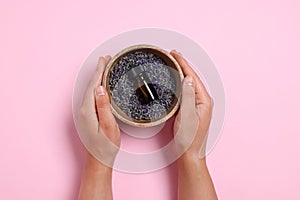 Woman holding bowl with lavender essential oil and flowers on pink background, top view