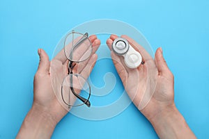 Woman holding case with contact lenses and glasses on light blue background, top view