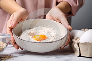 Woman holding ceramic bowl with flour and egg at white marble table in kitchen, closeup. Cooking oatmeal dough