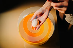 A woman is holding a chicken egg in her hand over a bowl. She prepares to cook it. The home cooking and the process of preparing