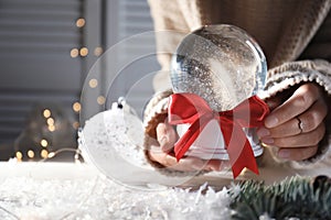 Woman holding Christmas snow globe with red bow on blurred background, closeup.