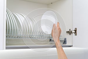 Woman holding clean and white plate near dishware in kitchen cabinet