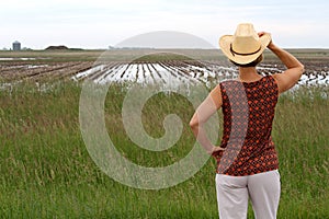 Woman holding cowboy hat looking at a farm field full of water.