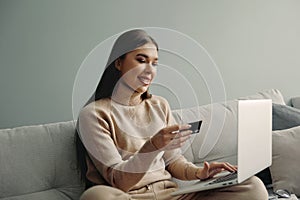 Woman is holding credit card and using laptop computer  sitting on couch at home,. Online shopping