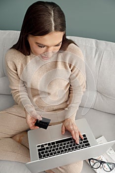 Woman holding credit card and using laptop computer sitting on couch at home. Online shopping concept.