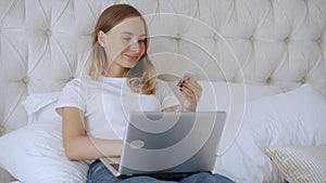 Woman holding credit card and using laptop, sitting on bed at home, young female customer shopping online in internet