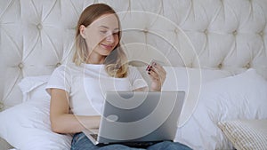 Woman holding credit card and using laptop, sitting on bed at home, young female customer shopping online in internet