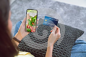 Woman holding credit card and using smartphone touching application screen for ordering salad online at sofa in the home.