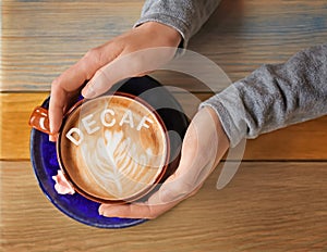 Woman holding cup of aromatic decaf coffee with foam at wooden table, top view