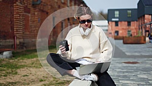 Woman holding cup of coffee and enjoying working on laptop while sitting outside