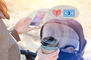 Woman holding cup of coffee to go and using smartphone during walk with baby carriage. Browsing social media, likes and messages.