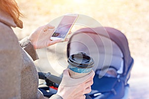 Woman holding cup of coffee to go and using smartphone during walk with baby carriage. Modern young business woman