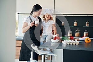 Woman is holding a cup with drink. Mother with her daughter are preparing food on the kitchen