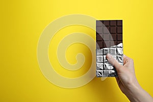 Woman holding delicious dark chocolate bar on yellow background, closeup. Space for text