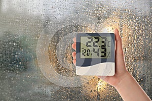 Woman holding digital hygrometer with thermometer near window on rainy day, closeup. Space for text