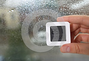 Woman holding digital hygrometer with thermometer near window on rainy day, closeup. Space for text