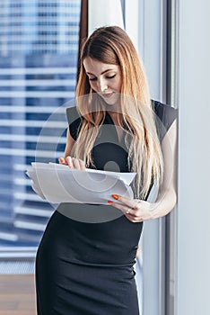 Woman holding documents, looking through papers, studying the report standing near window with view on skyscrapers