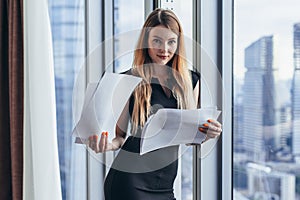 Woman holding documents, looking through papers, studying the report standing near window with view on skyscrapers