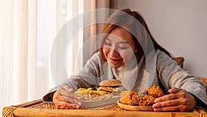 A woman holding and eating a hamburger, french fries and fried chicken on the table at home
