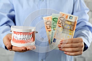 Woman holding educational dental typodont model and euro banknotes on light background, closeup. Expensive treatment