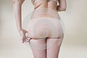 Woman holding fold of skin on buttocks, cellulite on female body, beige background