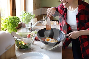 Woman holding frying pan with cooked eggs and vegetables at countertop in kitchen, closeup