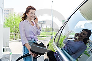 Woman holding fuel pump while refueling car with benzine and talking on smartphone