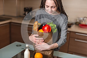 Woman holding full paper bag with products in hands on the background of the kitchen. Healthy and fresh organic food