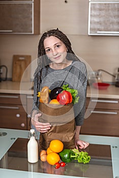 Woman holding full paper bag with products in hands on the background of the kitchen. Healthy and fresh organic food