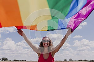 Woman holding the Gay Rainbow Flag over blue and cloudy sky outdoors. Happiness, freedom and love concept for same sex couples.