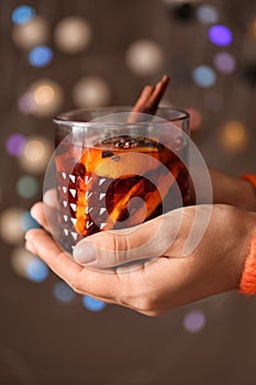 Woman holding glass of delicious mulled wine against blurred lights, closeup