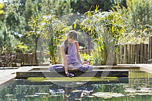 A woman holding a glass purple lotus while sitting in lotus pose on a tree trunk over the water