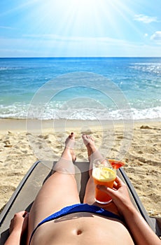 Woman holding in hand glass with drink and lying on a sun lounger on sandy beach.