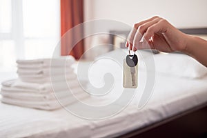 Woman holding in hand room key with blank label at hotel suite, copy space. Bedroom interior in blur