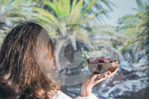 Woman holding in a hand royal dates fruit in a bowl of coconut in a palm tree grove