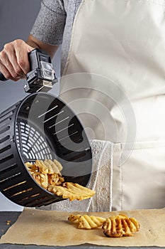 A woman is holding the handle of an air fryer oven basket with homemade fresh waffle potato fries inside