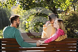 Woman holding hands with another man behind her boyfriend`s back on bench in park. Love triangle