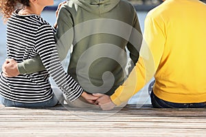 Woman holding hands with another man behind her boyfriend`s back on pier near river, closeup. Love triangle