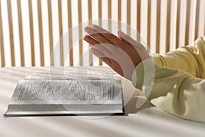 Woman holding hands clasped while praying over Bible at white table indoors, closeup