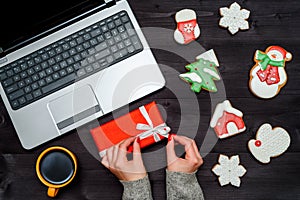 Woman holding in hands red Christmas present. Wrapping concept. Laptop computer, cup of coffee and gingerbread cookies.
