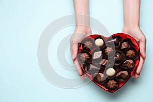 Woman holding heart shaped box with delicious chocolate candies on light blue background, top view. Space for text