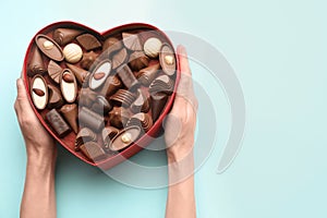 Woman holding heart shaped box with delicious chocolate candies on light blue background, top view. Space for text