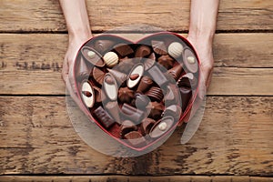 Woman holding heart shaped box with delicious chocolate candies at wooden table, top view